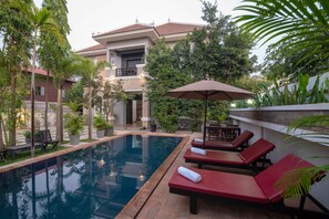 Un espace piscine avec des chaises longues et une table, entouré de verdure et bordé d’un bâtiment doté d’un balcon.