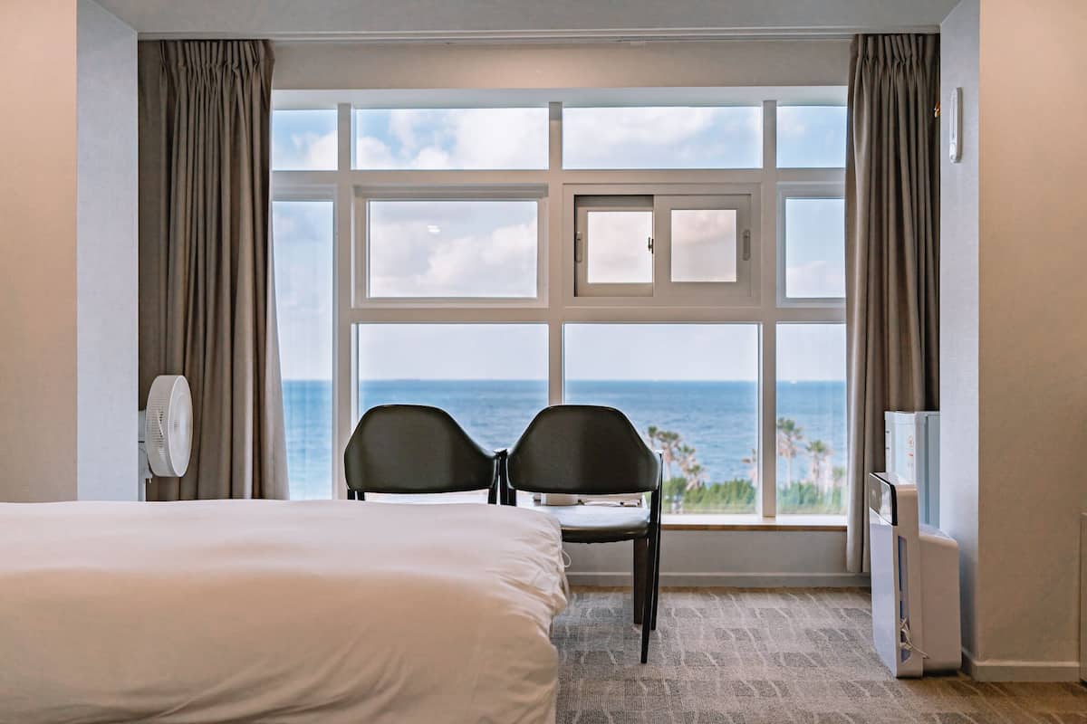 A hotel room with a bed, two chairs, a large window with a view of the ocean, and a wall-mounted air conditioner.
