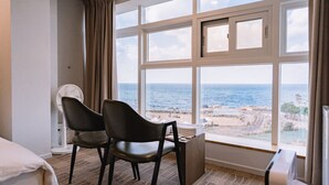 Quarto de hotel com uma grande janela com vista para o mar e a praia. Há uma cama, duas cadeiras, uma escrivaninha com luminária e um ventilador.