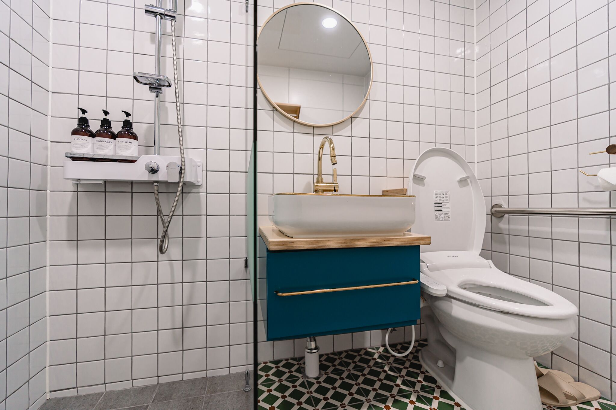 Un baño moderno con pared de azulejos blancos, un espejo redondo, un lavabo blanco con grifo dorado, un mueble azul y un inodoro blanco.