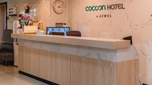 Bancada de recepção de hotel com um relógio, um computador e uma placa que diz "coccon hotel in AEWOL".
