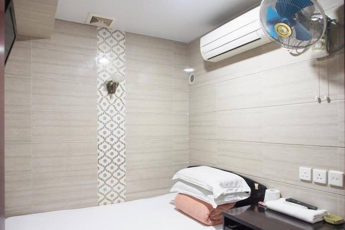 Una habitación de hotel con cama, mesita de noche, aire acondicionado de pared y ventilador de pared.