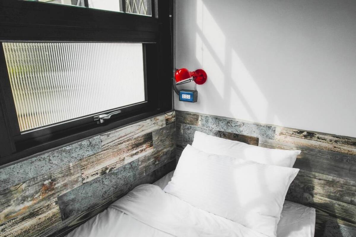 Una cama con ropa blanca, cabecera de madera, ventana con persianas y una lámpara roja en la pared.