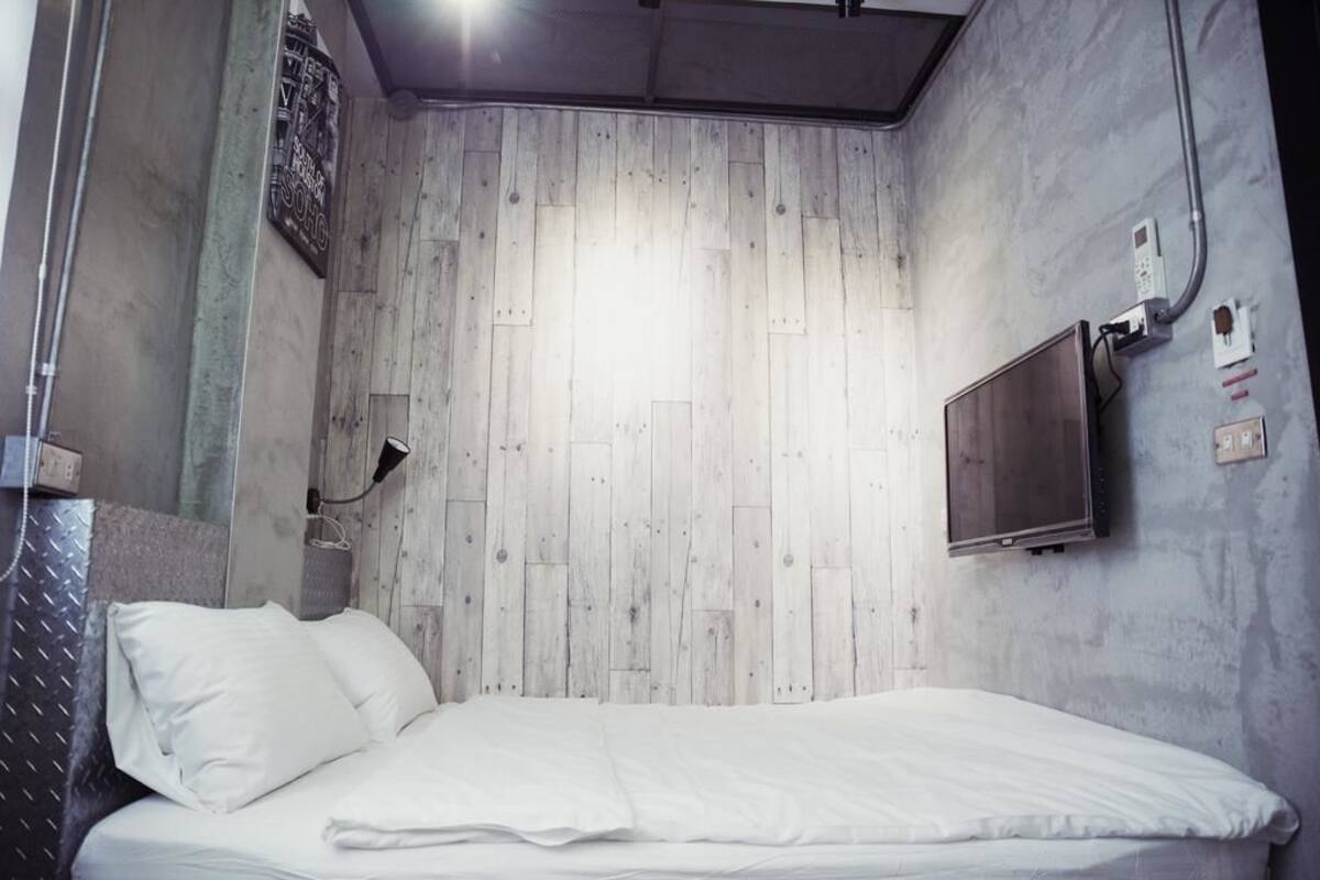 Un dormitorio con panel de madera en la pared, una cama con ropa de cama blanca, un televisor montado y un enchufe en la pared.