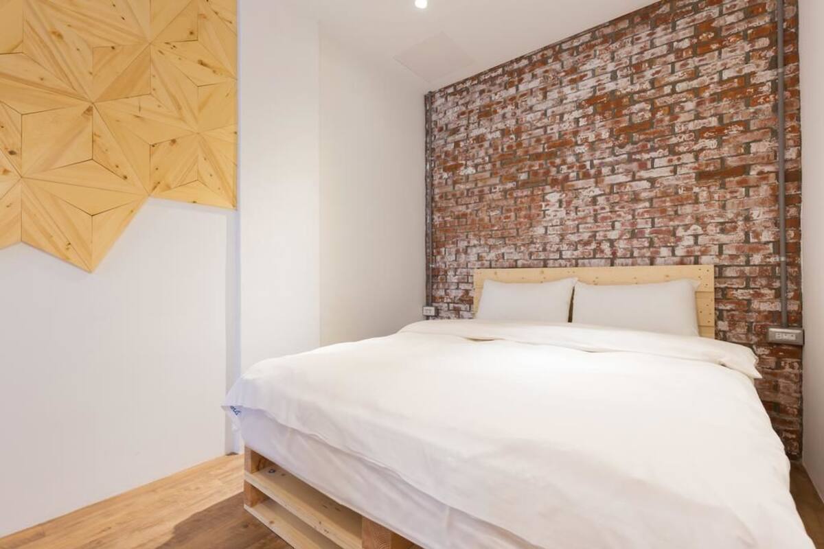 Un dormitorio con un cabecero de ladrillo, una obra de arte mural de madera con diseño geométrico y una colcha blanca.