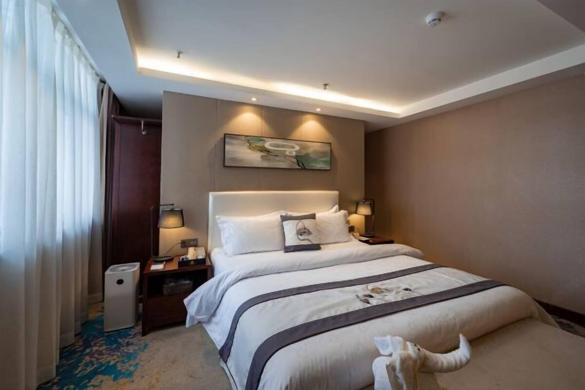 A hotel room with a large bed, bedside lamps, a nightstand, a trash bin, and a ceiling-mounted security camera.