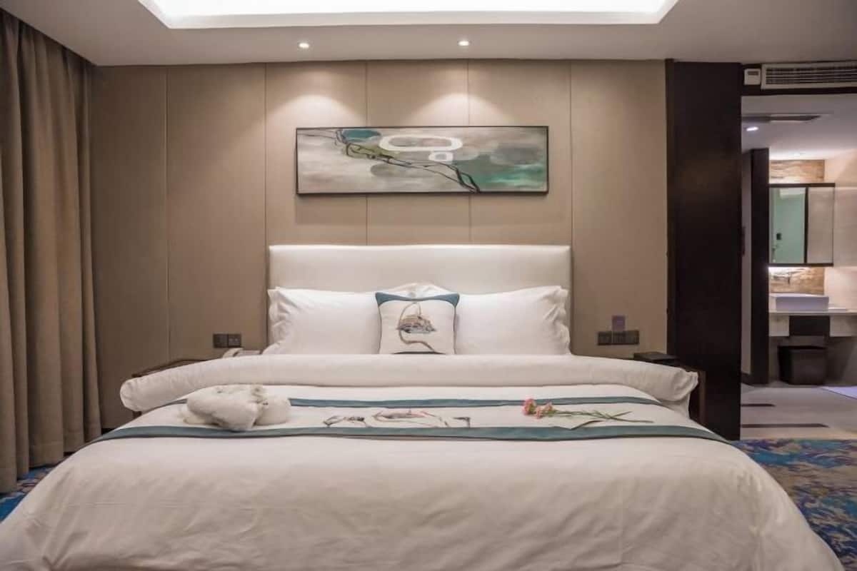 A modern hotel room with a large bed, a headboard with a decorative pillow, a painting above the bed, and a view of the bathroom through an open door.