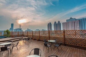 Eine Dachterrasse mit Tischen und Stühlen, ein Holzboden und Blick auf eine Stadtlandschaft mit Gebäuden und einem Turm.