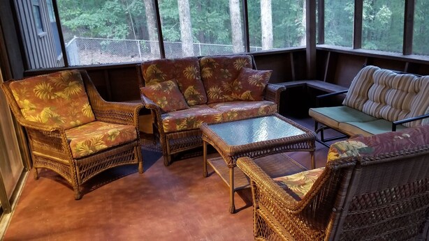 A sunroom with wicker furniture, a glass coffee table, and a view of the forest through large windows. - Canterbury Cabin (Hot Springs)