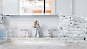 A bathroom with a white marble sink, two chrome faucets, and a stack of white towels with a leaf pattern.