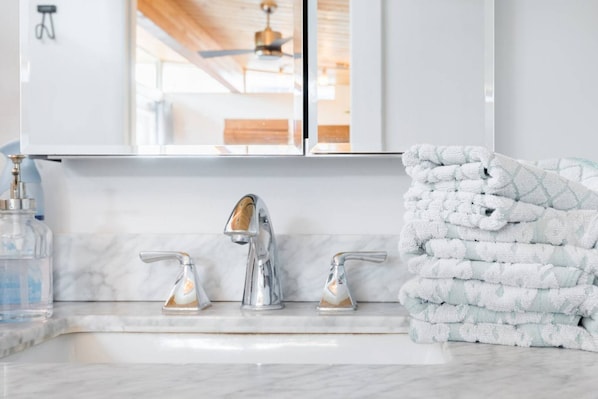 A bathroom with a white marble sink, two chrome faucets, and a stack of white towels with a leaf pattern.
