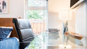 A modern hotel room with a glass table, a leather chair, a bed with blue bedding, a window with a view of greenery, and a lamp.