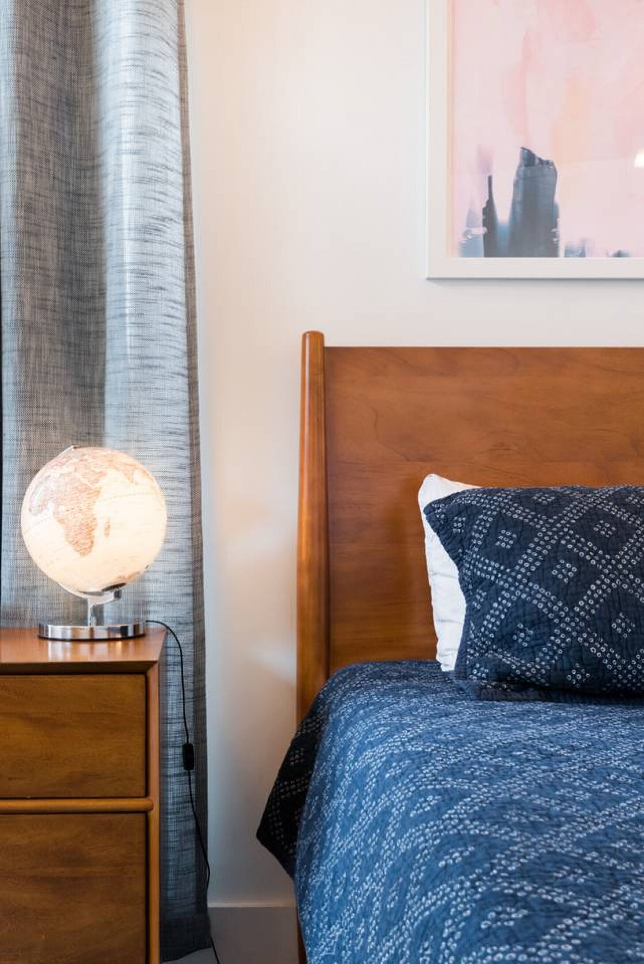 Una cama bien hecha con una colcha azul estampada y una sola almohada, una mesita de noche de madera con una lámpara de globo, y un cuadro en la pared.