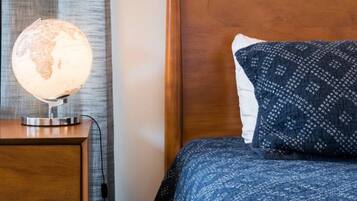 Una cama bien hecha con una colcha azul estampada y una sola almohada, una mesita de noche de madera con una lámpara de globo, y un cuadro en la pared.