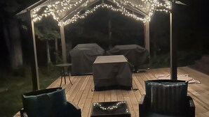 Een terras buiten met een overdekte zithoek, een vuurkorf en lichtslingers.