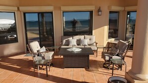 A sunlit patio with a sofa, chairs, and a table, featuring large windows with a view of the landscape.