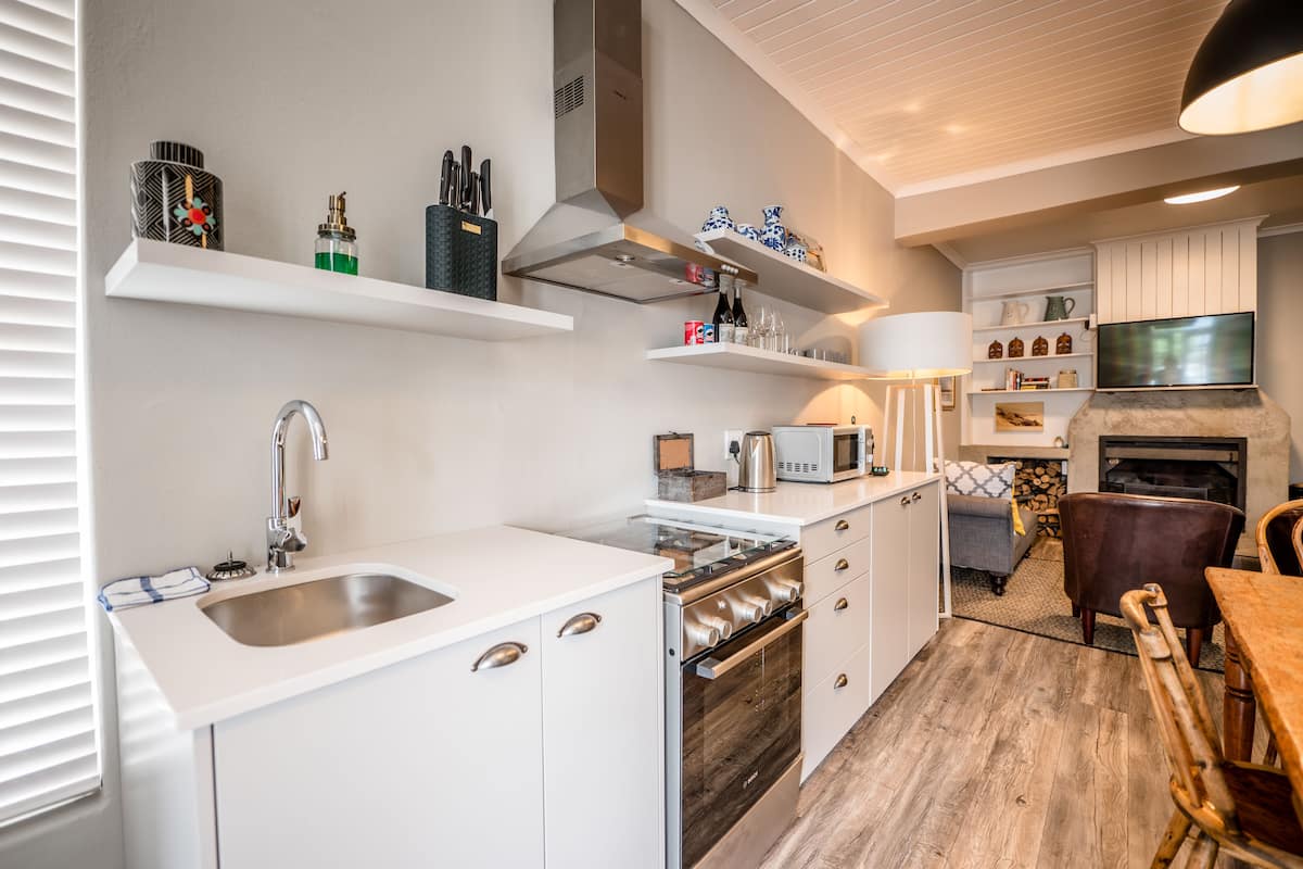 A modern kitchen with white cabinetry, stainless steel appliances, and a built-in oven. There is a dining area with a wooden table and chairs, and a fireplace in the background.
