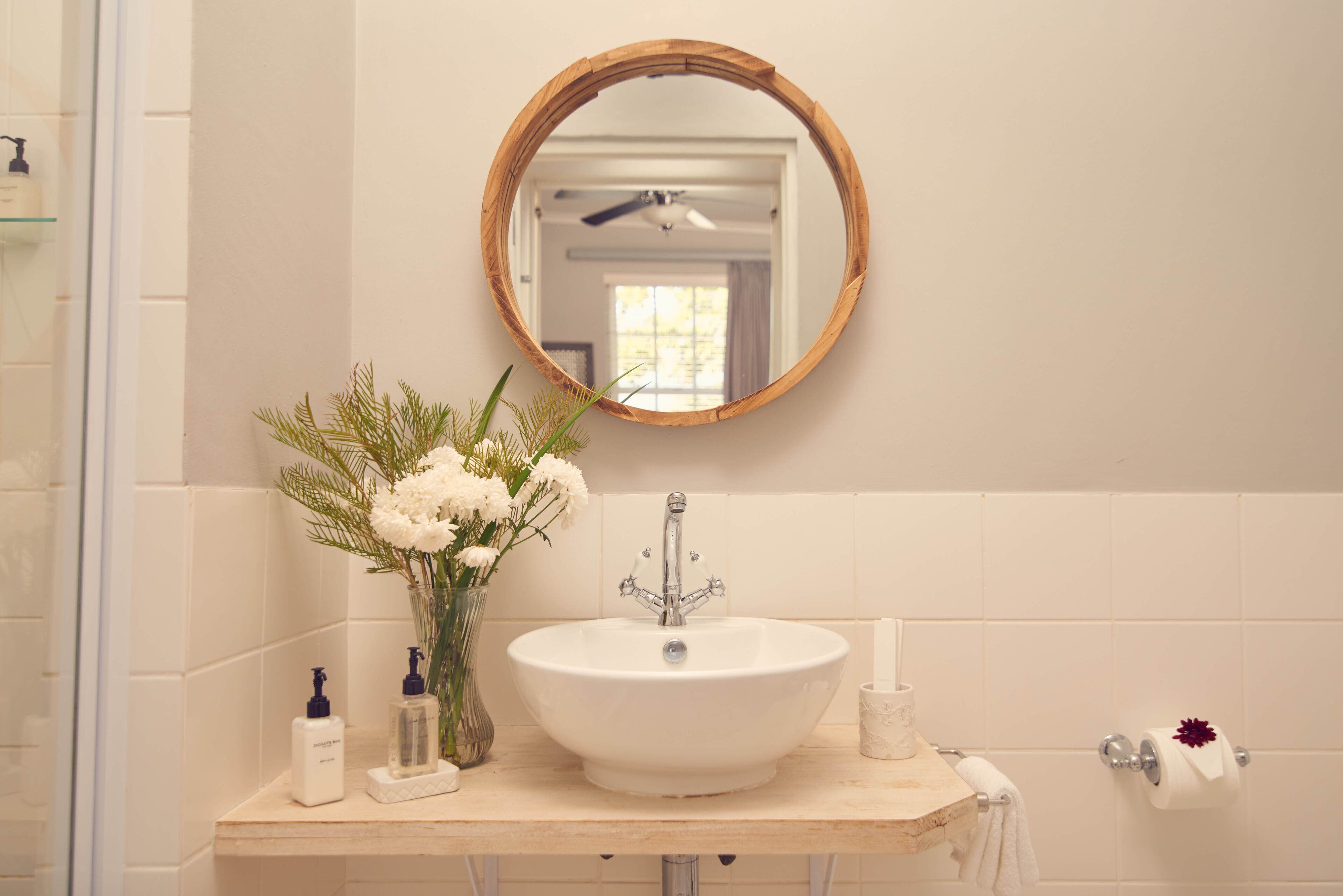 Un baño con un espejo redondo de marco de madera, un lavamanos blanco y un florero con flores blancas.