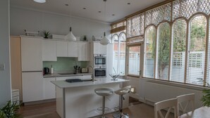 A modern kitchen with white cabinetry, a central island with bar stools, a microwave, oven, and a window with blinds. - Toothbrush Apts - Ipswich Fonnereau Road (Ipswich)