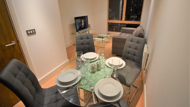 A dining area with a glass table set for four, surrounded by chairs, a television, and a window with a city view. - Toothbrush Apartments - Ipswich Quayside (Ipswich)