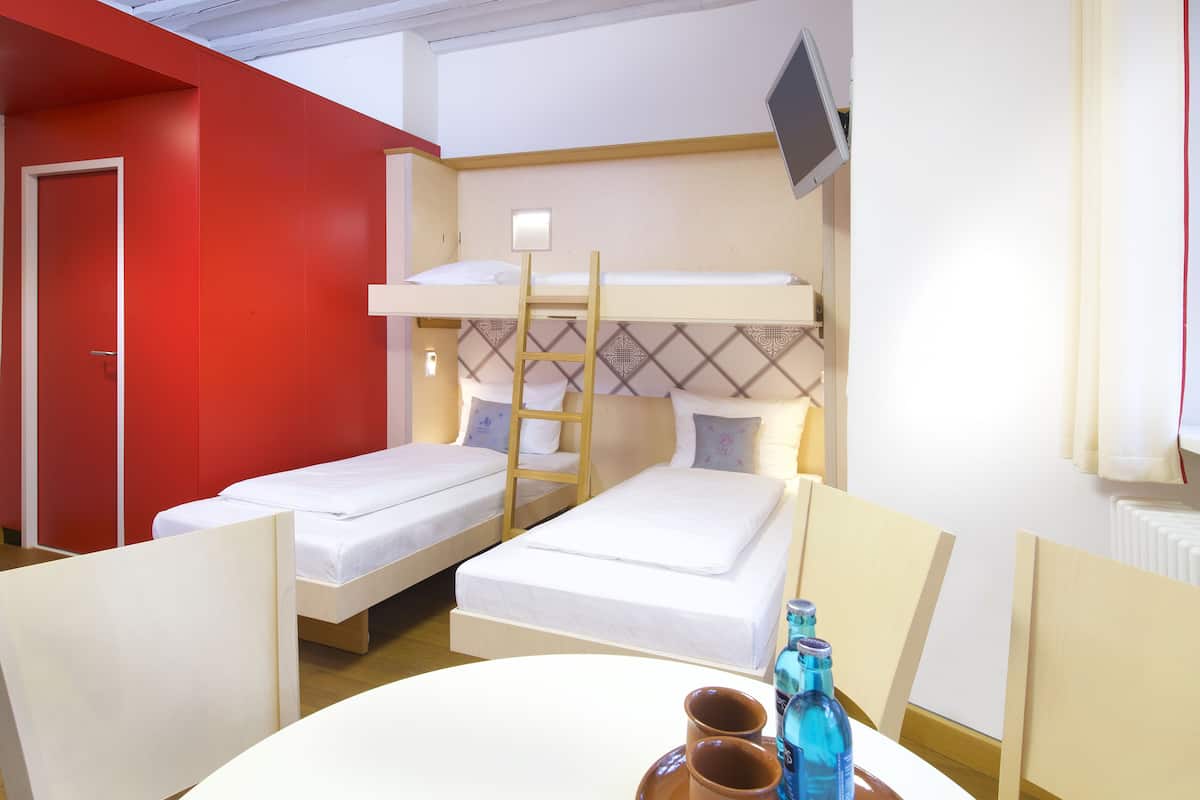 A room with two single beds, a bunk bed, a table with two cups, and a TV mounted on the wall.