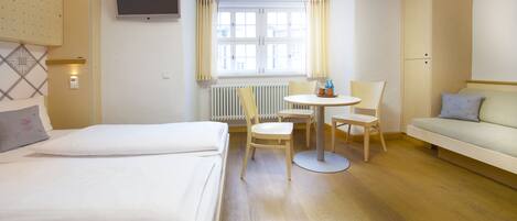 A hotel room with two beds, a round table with chairs, a sofa, a TV mounted on the wall, and a window with curtains.