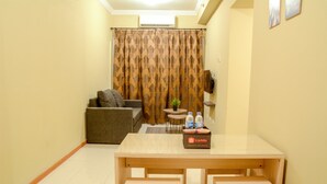 A small, well-lit room with a wooden table, two stools, a grey sofa, a small round table with a plant, and a curtain with a pattern. - Big 2 BR (76 sqm) Apartment Grand Palace/Pallazo Kemayoran (Jakarta)