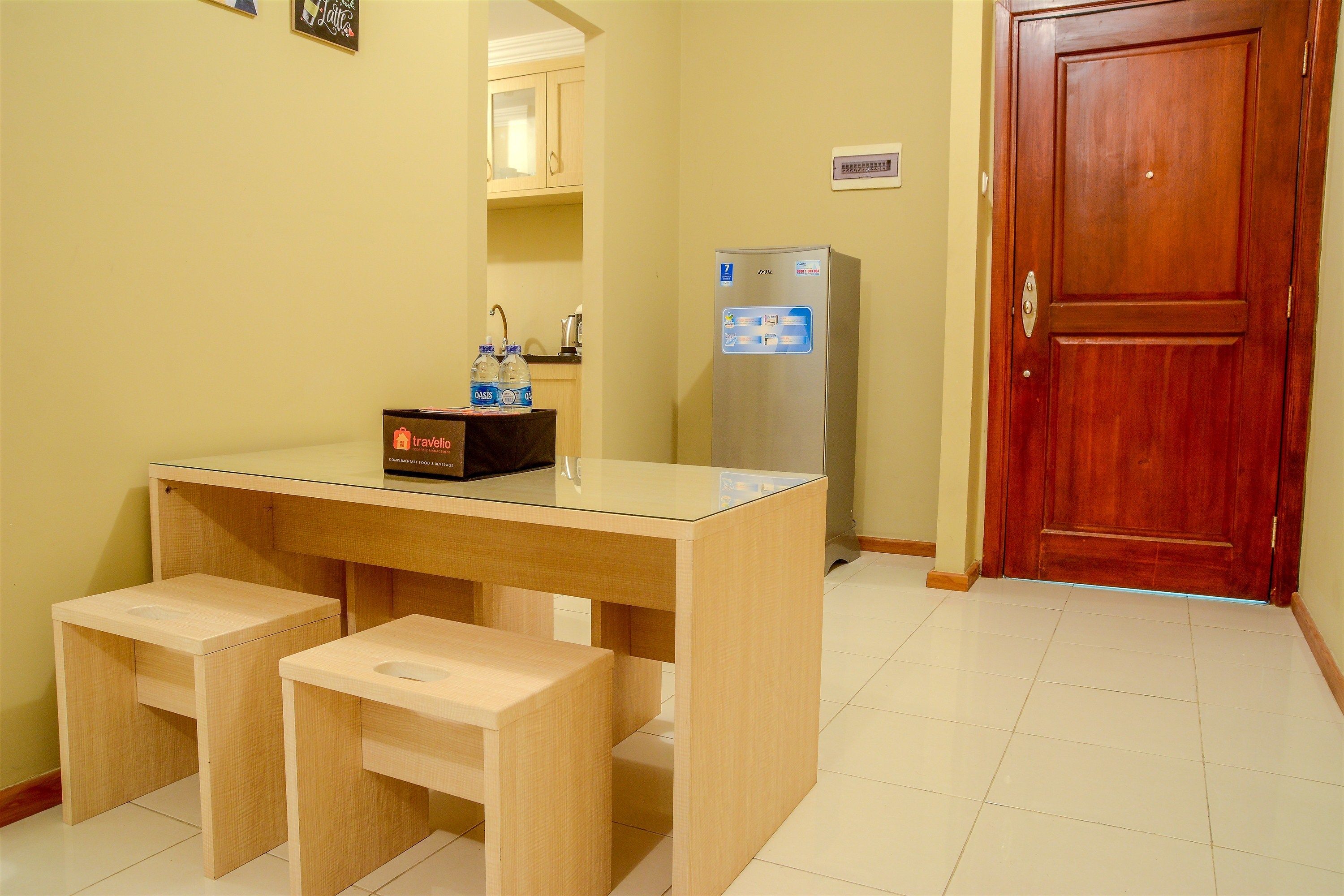 A compact kitchenette with a wooden table, two stools, a refrigerator, and a wooden door.