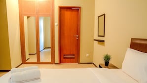 A hotel room with a wooden door, a bed with white linens, a bedside table with a plant, and a mirror on the wall. - Best of the Best 3BR Apartment Grand Palace/Pallazo Kemayoran (Jakarta)