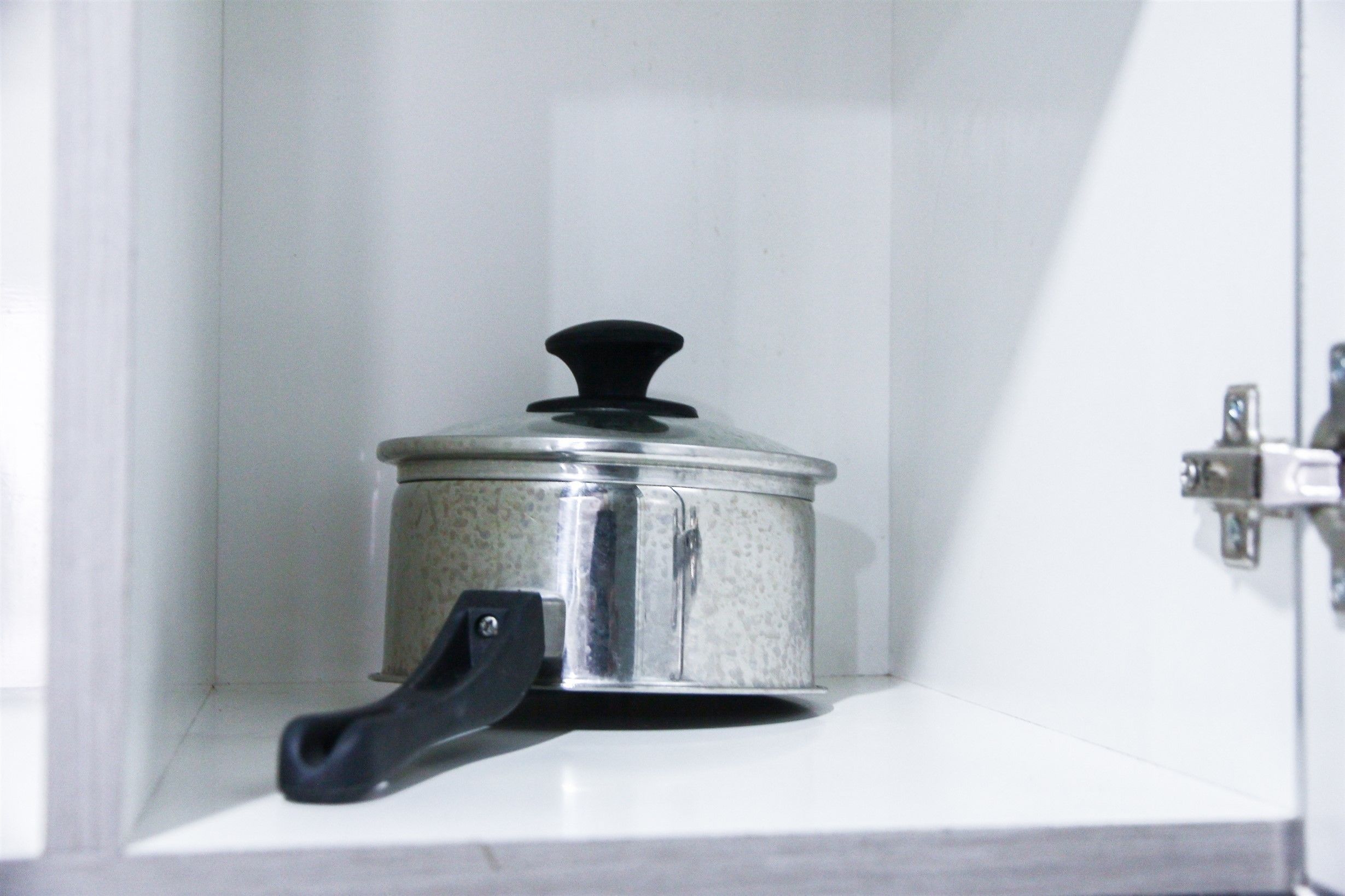 A stainless steel pot with a black handle on a white shelf in a kitchen cabinet.