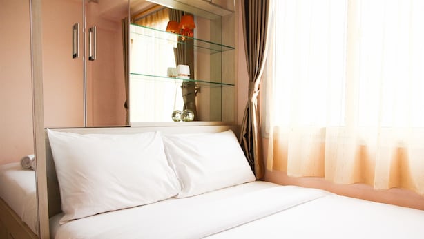 A neatly made bed with white linens, a glass shelf with a lamp and decorative items, and a mirror reflecting the room's interior. - Best Price 2BR Mutiara Bekasi Apartment (Bekasi)
