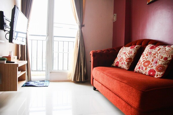 Ein Wohnzimmer mit einem roten Sofa, floralen Kissen, einem Holztisch und Blick auf einen Balkon durch ein mit Vorhängen drapiertes Fenster.