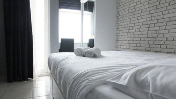 A hotel room with a white brick wall, a neatly made bed with white bedding, and a view of the outdoors through a window. - Modern and Comfortable Studio Apartment Springlake Summarecon near Summarecon (Bekasi)