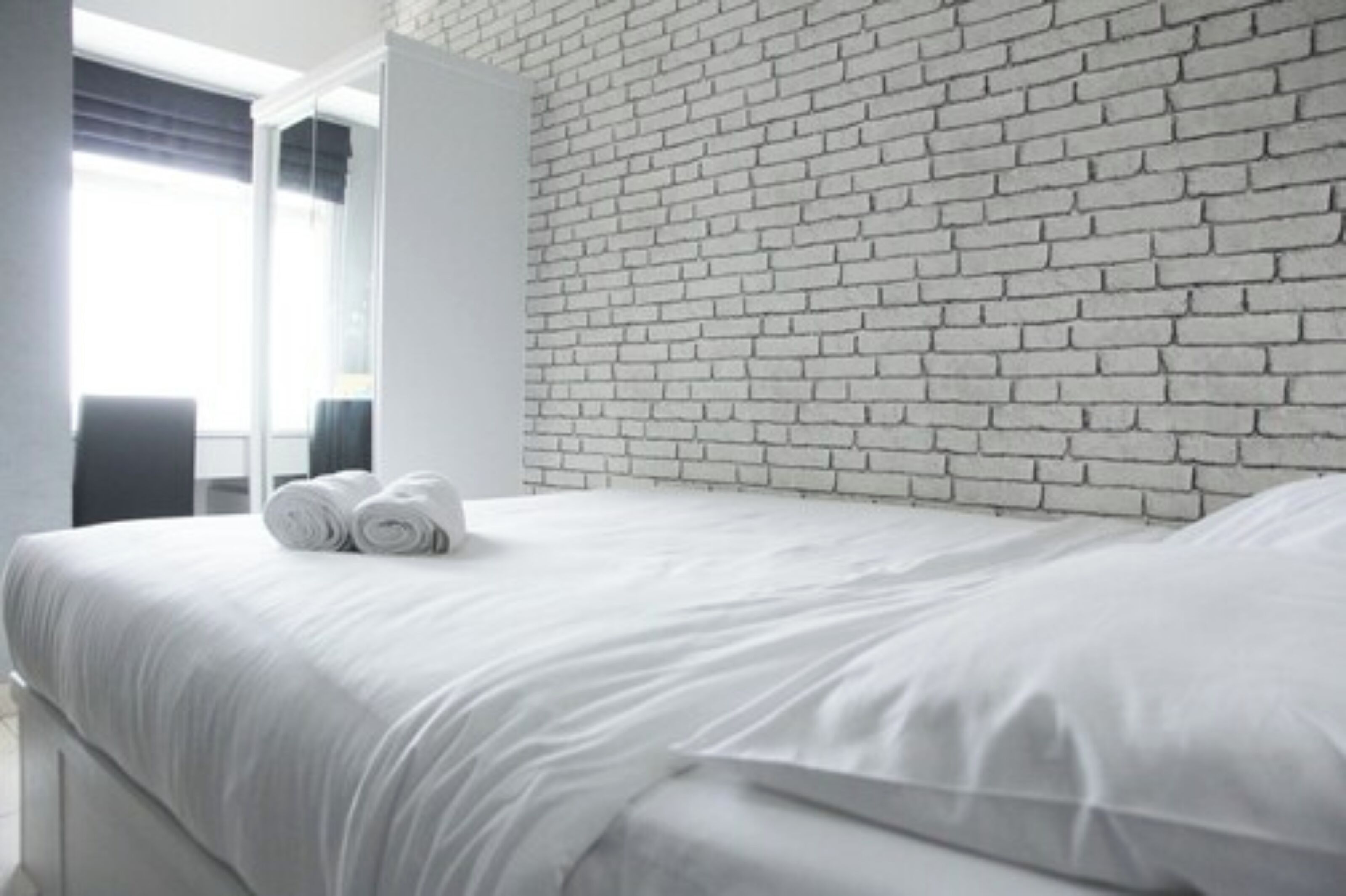Une chambre d’hôtel moderne avec un mur en briques blanches, un lit bien fait avec du linge de lit blanc et une fenêtre avec des stores.