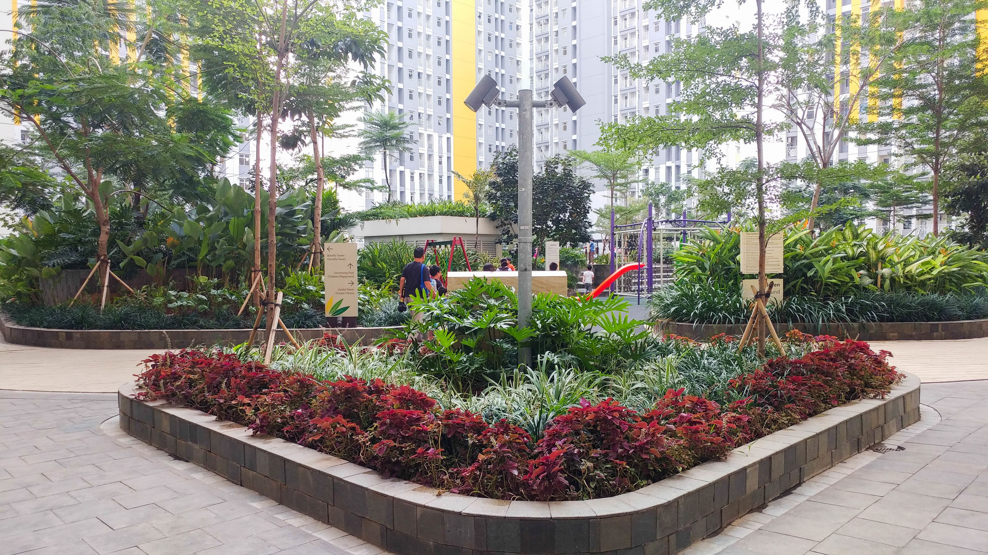 Un espace urbain aménagé avec un jardin central, une aire de jeux et un grand immeuble d’habitation en arrière-plan.