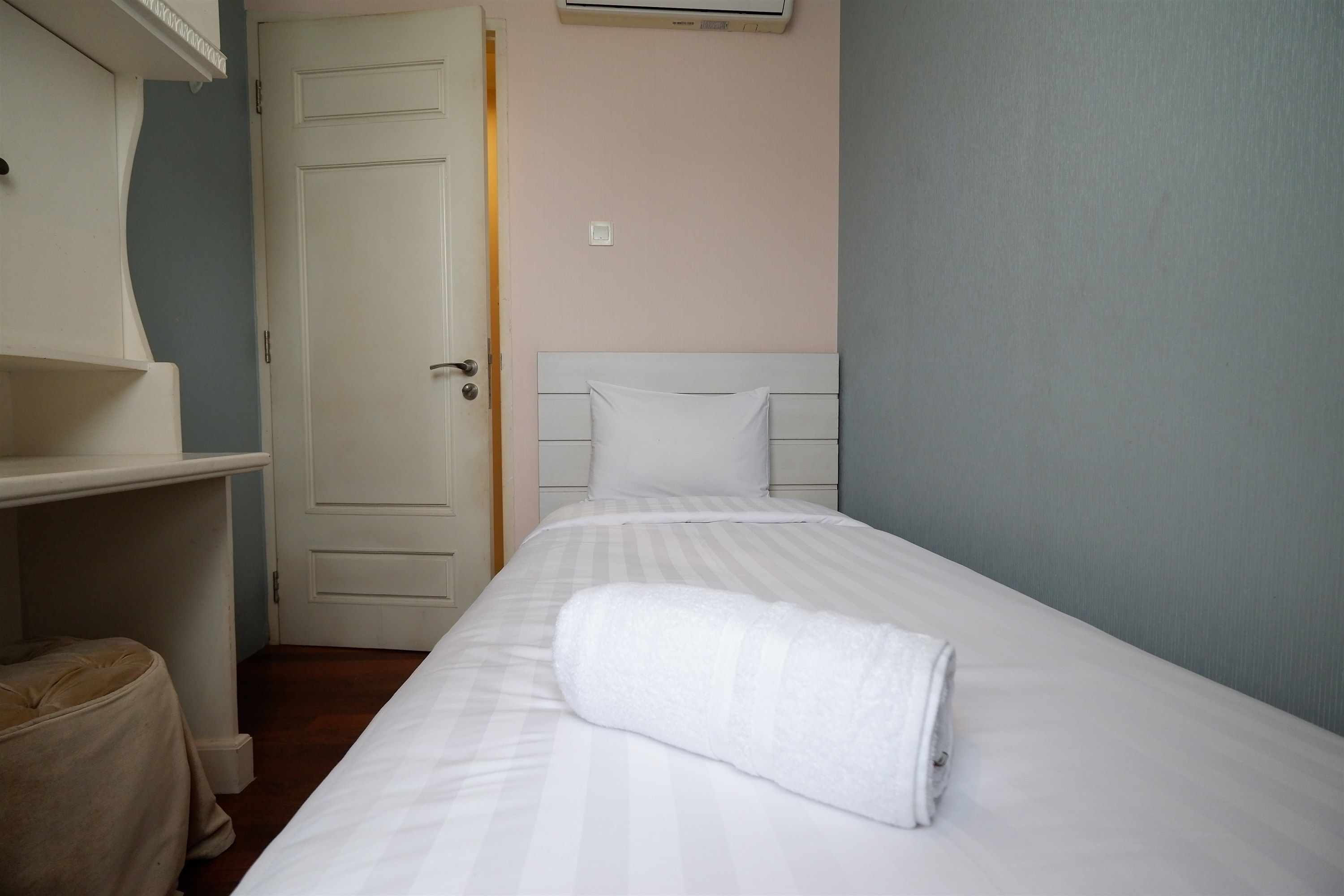 A neatly made bed with a rolled white towel, a white headboard, and a closed door in a room with light-colored walls.