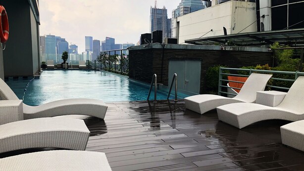 A rooftop pool area with white wicker lounge chairs, a metal ladder, and a city skyline view in the background.