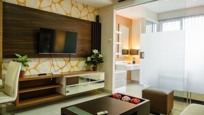 A modern living room with a flat-screen TV, a wooden entertainment unit, a coffee table with flowers, a sofa, and a glass partition leading to a kitchen area.
