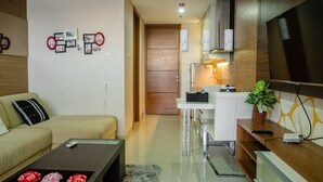 A modern living room with a sofa, coffee table with flowers, a TV, and a kitchen area with a fridge and microwave.