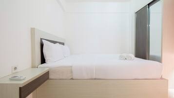Una cama bien tendida con sábanas blancas y dos toallas enrolladas, una mesita de noche con un libro, y un ventanal con vistas al exterior.