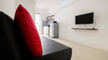 Una sala de estar moderna con un sofá negro, un cojín rojo, un televisor de pantalla plana montado en la pared y una zona de cocina al fondo.