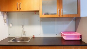 A kitchen with wooden cabinets, a stainless steel sink, a pink plastic container, and a white wall with a phone and a small red object. - Pool View Studio Apartment Tamansari Sudirman (Jakarta)