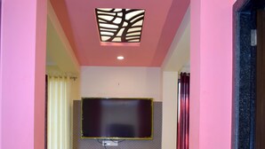A hotel room with a pink wall, a flat-screen TV mounted on the wall, a small table with a lamp, and a trash bin. - Hotel Shree Pratham Milan (Nathdwara)
