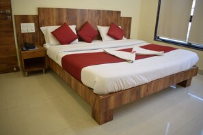 A large bed with a wooden frame, white bedding, and red pillows. There is a wooden nightstand with a phone and a power outlet. - Hotel Shree Pratham Milan (Nathdwara)