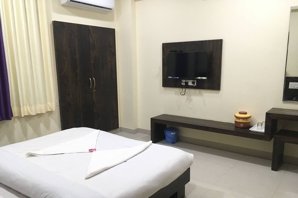 Quarto de hotel com cama, guarda-roupa, televisão e uma pequena mesa com chaleira e copos.