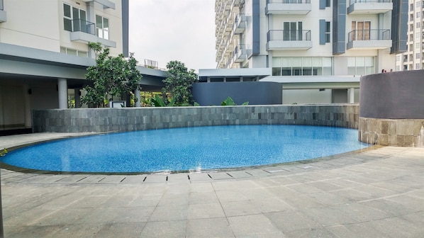 Una piscina con plataforma de baldosas, rodeada por un bajo muro de piedra y edificios modernos al fondo.
