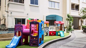 A children's play area with various colorful play structures, including slides and climbing frames, in front of a building with balconies and windows. - 2BR Apartment with City View at Mediterania Marina Residences (Jakarta)