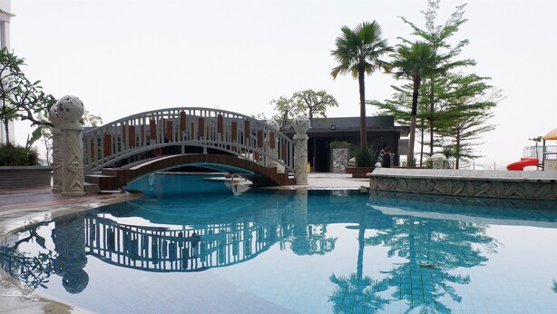 A swimming pool with a decorative bridge, palm trees, and a building in the background. - Close to Pool Studio Apartment Sky Terrace at Daan Mogot Baru (Jakarta)