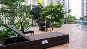 A wicker lounge chair and table set in a courtyard with a pool, surrounded by greenery and high-rise buildings.