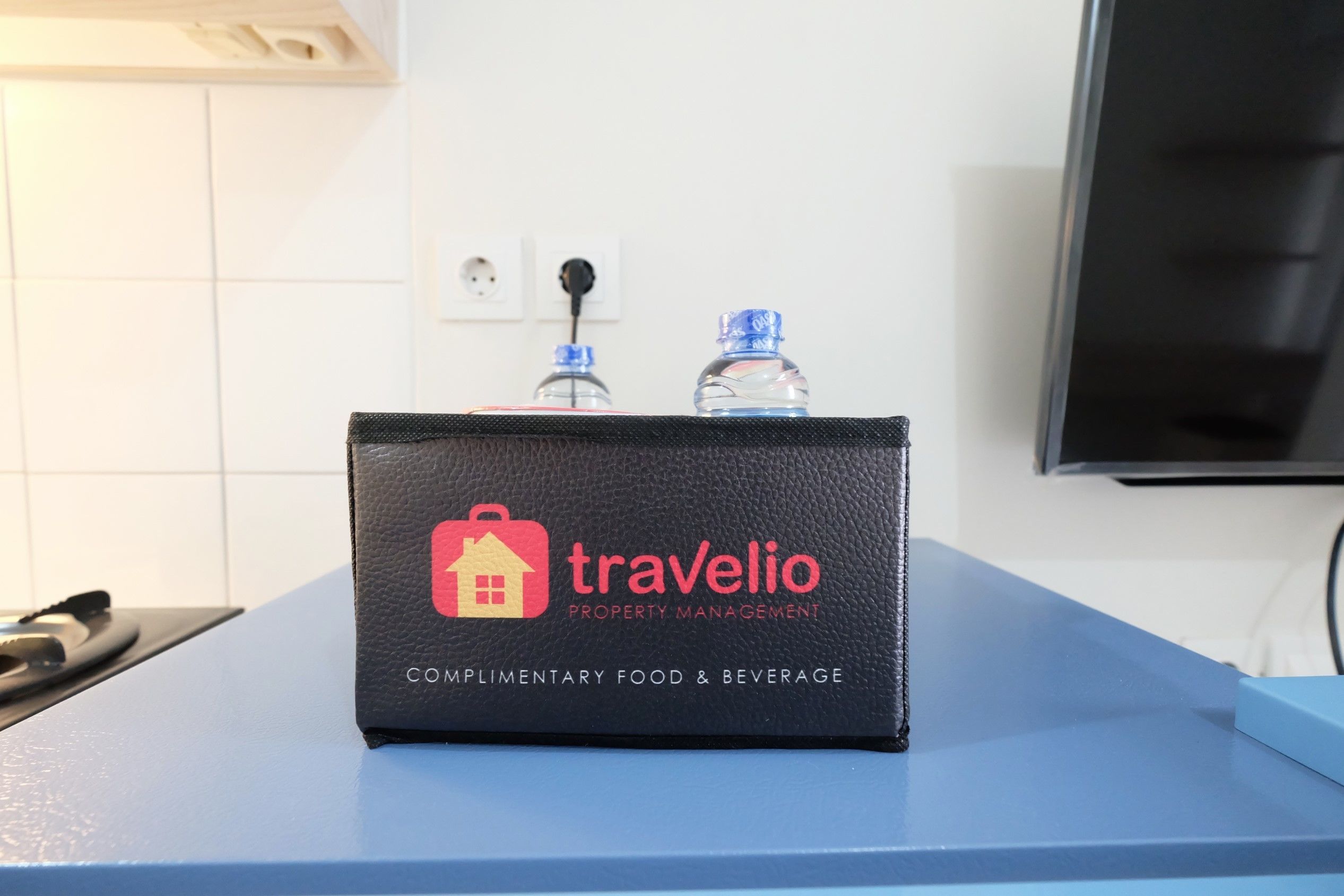 A black leather bag with the logo "Travelio Property Management" and the text "Complimentary Food & Beverage" on a blue counter.